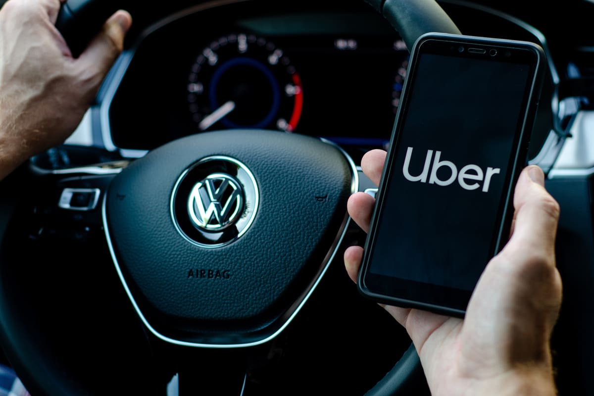 main posée sur le volant d'une voiture Volkswagen et l'autre main tenant un smartphone avec l'écran affichant Uber 